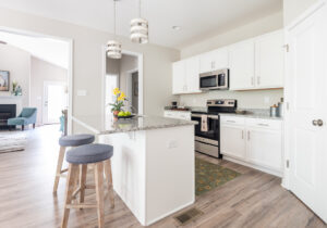 A modern kitchen with white cabinets, granite countertops, stainless steel appliances, an island with two stools, and pendant lights. An adjacent living room is partially visible in the background.
