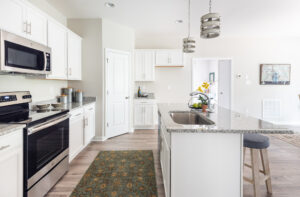 A modern kitchen with white cabinets, stainless steel appliances, granite countertops, a central island with a sink, and hanging pendant lights. There's a green rug on the floor and a potted orchid on the island.