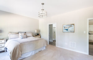 A modern bedroom with a neatly made bed, wall art, a bedside table with a lamp, and an open door leading to an en-suite bathroom and another room. The floor is carpeted, and a ceiling light fixture hangs above.
