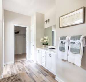 Bright bathroom with white cabinetry, wood-style flooring, a single sink, framed starfish artwork, hanging towels, and a doorway leading to an empty walk-in closet.