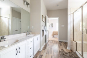 A modern bathroom with a double sink vanity, white cabinets, a glass shower, and wood laminate flooring. A doorway opens to a bedroom with a bed in the background.
