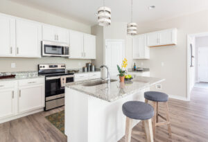 A modern kitchen with white cabinets, stainless steel appliances, a granite countertop island, two gray cushioned stools, and pendant lights above the island.