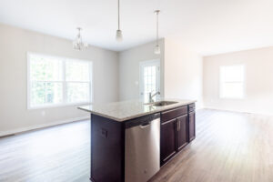 A spacious, modern kitchen with a central island, granite countertop, sink, stainless steel dishwasher, pendant lights, and large windows allowing natural light to fill the room.