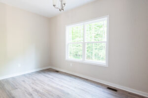 The spacious room in Rowland features light wood flooring and beige walls. A large window reveals the lush greenery of Poplar Village, while a sleek ceiling light fixture adds a modern touch.