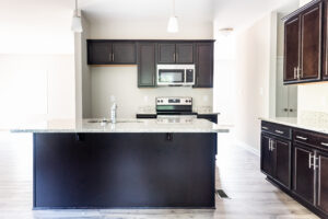 Modern kitchen with dark wood cabinets, granite countertops, stainless steel appliances, and an island with sink and faucet. Two pendant lights hang above the island.
