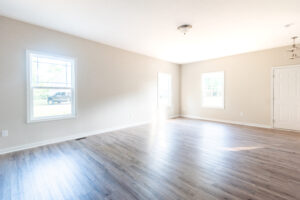 A bright, empty room with light gray walls, large windows, a wooden floor, and a ceiling light fixture. A door is situated on the right side.