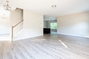 An empty, well-lit room with light wood flooring, light-colored walls, a staircase on the left, and a glimpse of a kitchen area and windows in the background.