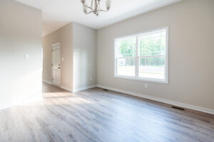 A bright, empty room with light-colored wooden flooring, beige walls, a white door, a large window, and a ceiling light fixture. Natural light streams in from the window.