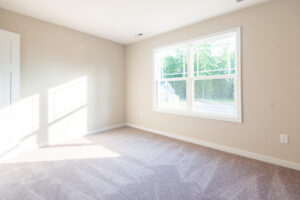 A bright, empty room with beige walls, a large window letting in natural light, and light gray carpeted flooring.