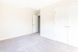 An empty room with beige walls, a closed double-door closet, a single open door, and carpeted floors.