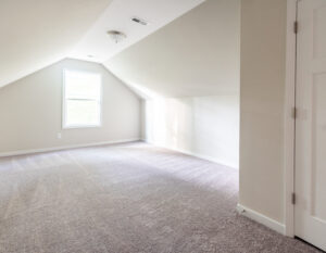 A clean, empty attic room with angled ceilings, a single window, beige walls, and carpeted floor, illuminated by natural light.
