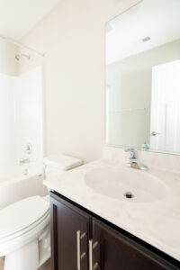 A bathroom featuring a white toilet, a sink with a marble countertop, and a large mirror. The shower area includes a curtain rod, and the walls are painted light beige.