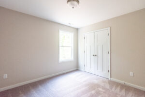 The empty room in Poplar Village features beige walls, a window, a closed white closet, and light gray carpet.