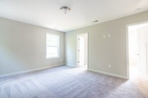 A brightly lit empty room with grey carpet, a window, a closed door on the right wall, and a partially open door on the back wall leading to another room.