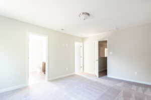 A bright, empty bedroom with clean carpet, beige walls, two open doorways leading to an adjoining bathroom and hallway, and a ceiling light fixture.