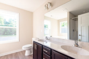 A bathroom with a large mirror over a double sink vanity. There is a toilet near a window, and a shower stall is partially visible in the background. The room is illuminated by natural light.