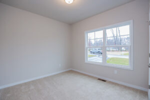 Empty room with beige walls, carpeted floor, and a large window overlooking Poplar Villages suburban street.