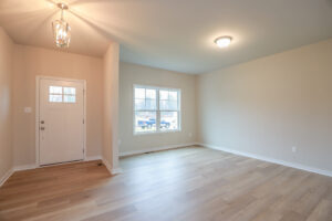 Empty room in Poplar Village, Lot 32, with wooden flooring, cream walls, a window, and a white front door illuminated by lights.