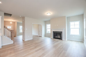 Spacious living room in Poplar Village, Lot 32, with light wood floor, fireplace, large windows; adjacent room and staircase visible.
