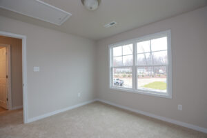 Empty room in Lot 32 with beige carpet, white walls, a large window, and open door to the left in Poplar Village.