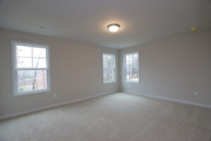 Empty beige carpeted room with three windows, light gray walls, and a ceiling light in Poplar Villages Lot 32.
