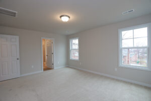An empty, carpeted room in Lot 32, featuring two windows and a door to a bathroom in Poplar Village.