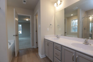 Modern Lot 32 bathroom with double sinks, large mirror, and wood flooring, leading to a carpeted Poplar Village room.