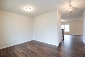 Empty room with wooden floors, white walls, and ceiling lights, leading to a naturally lit hallway in Poplar Village Lot 31.
