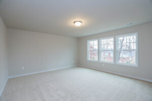 Empty room with beige carpet, white walls, ceiling light, and large windows showing a view of trees in Poplar Village.