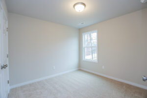 Empty room in Poplar Village with light beige walls, carpeted floor, and a window on the right. Ceiling light fixture is on.