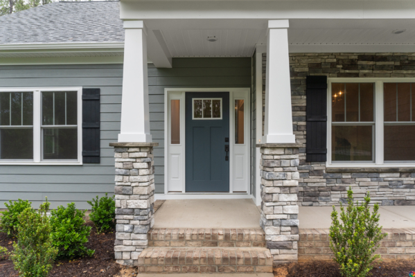 The front porch of this Scotchtown Estates home features a blue door, stone pillars, and gray siding, all beautifully complemented by the surrounding small shrubs.