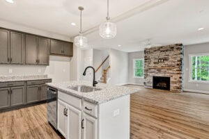 Modern kitchen with gray cabinets, island sink, and open living area featuring a stone fireplace and wood floors.