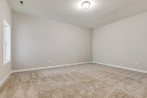 Empty carpeted room with beige walls, white trim, a ceiling light, and a window on the left wall.