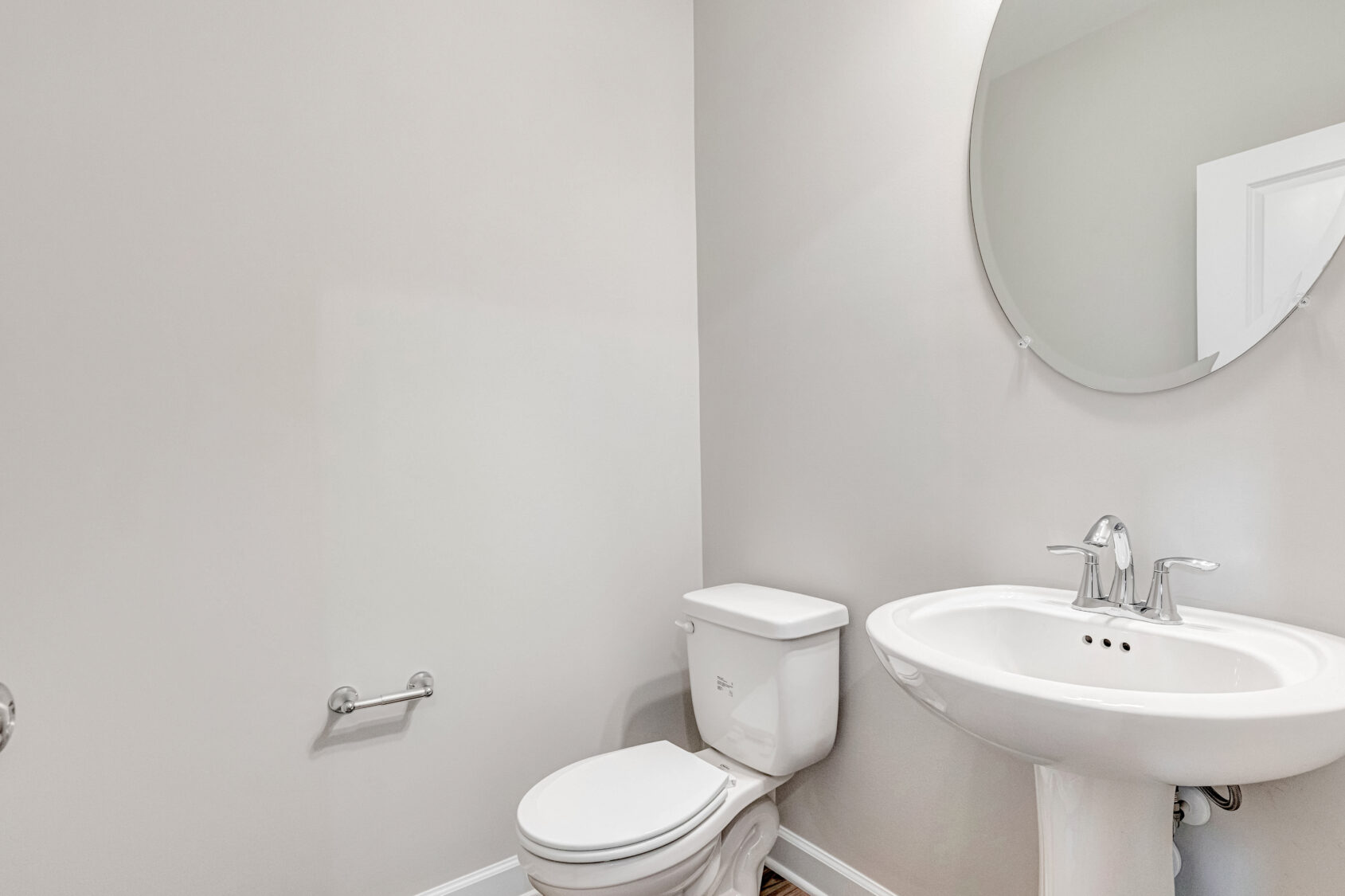 White toilet and pedestal sink in a small bathroom with light gray walls and a round mirror above the sink.