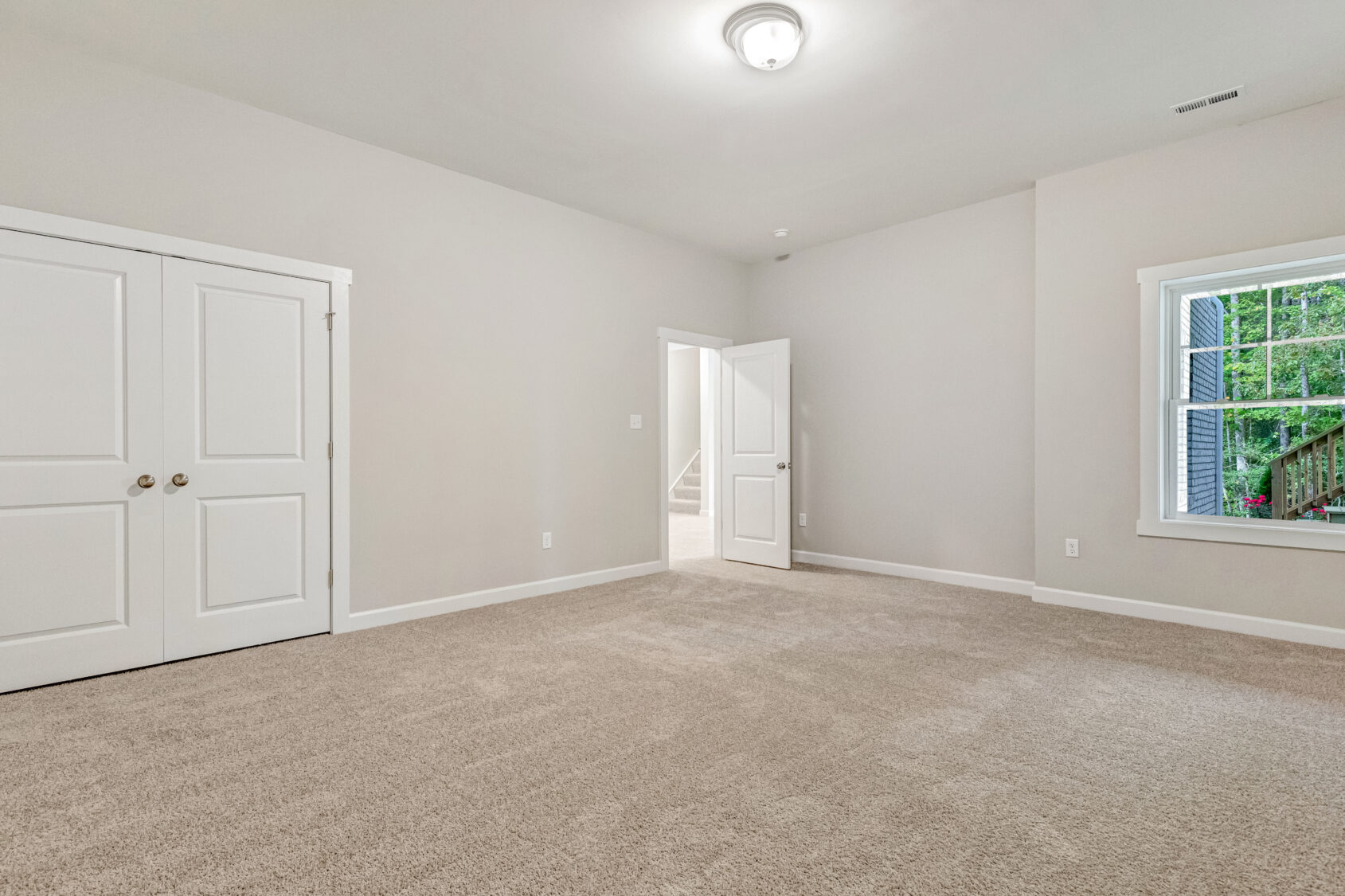 Empty carpeted room with beige walls, white trim, double closet doors, open door, and window with outdoor view.