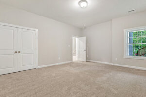 Empty carpeted room with beige walls, white trim, double closet doors, open door, and window with outdoor view.