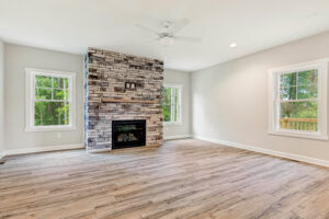 Bright living room with a stone fireplace, wood floors, ceiling fan, and large windows with outdoor views.