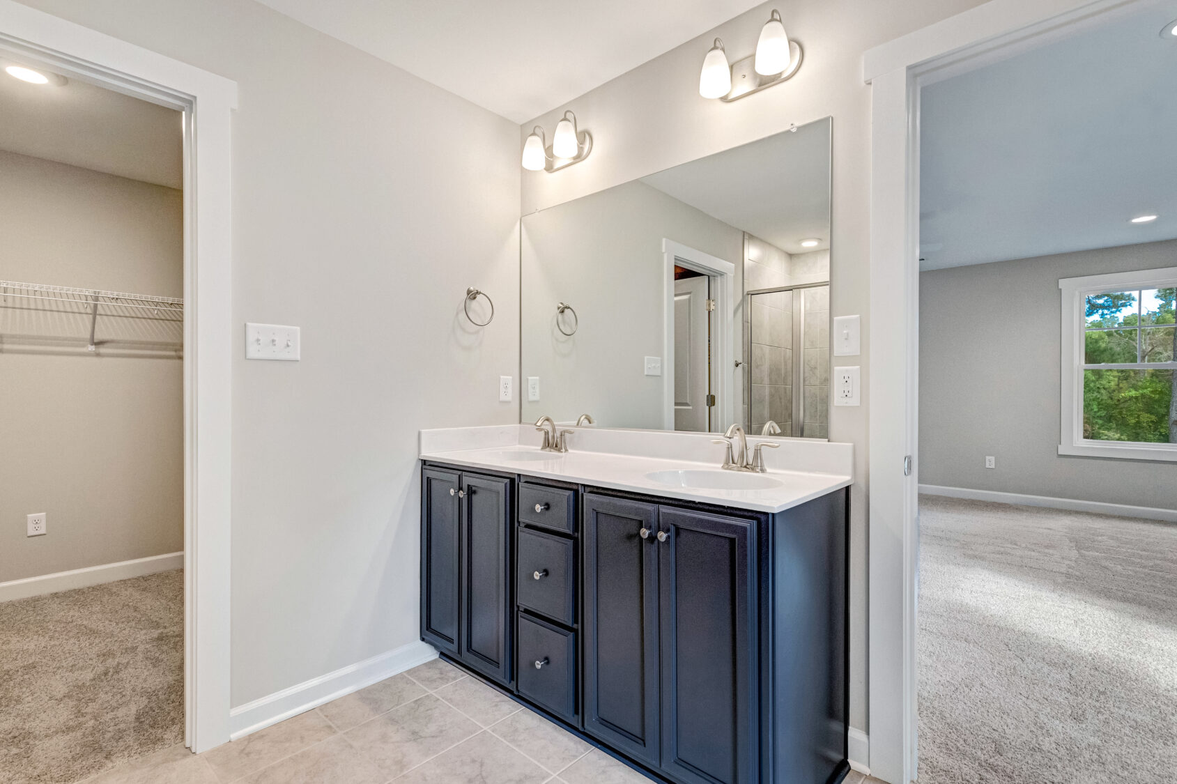 Modern bathroom with double sink vanity, large mirror, and view into a carpeted bedroom and walk-in closet.