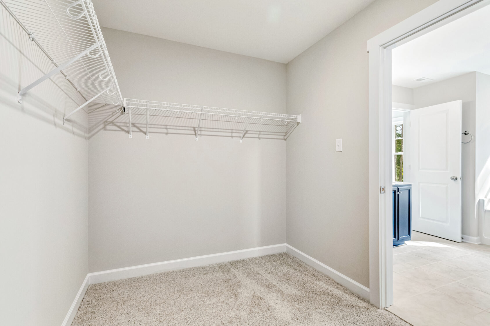 Empty walk-in closet with beige carpet, white wire shelves, and an open door leading to a bright bathroom.