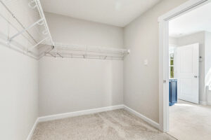 Empty walk-in closet with beige carpet, white wire shelves, and an open door leading to a bright bathroom.