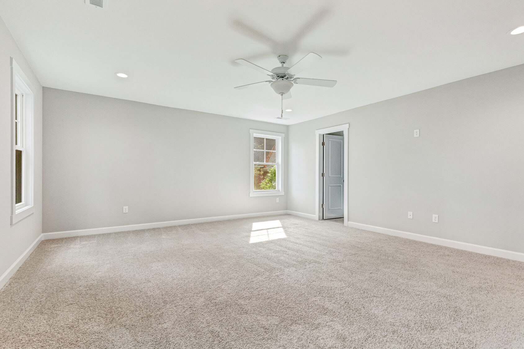 Empty, carpeted room with light gray walls, a ceiling fan, windows, and a door, lit by natural light.