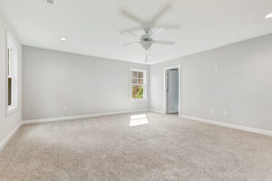 Empty, carpeted room with light gray walls, a ceiling fan, windows, and a door, lit by natural light.