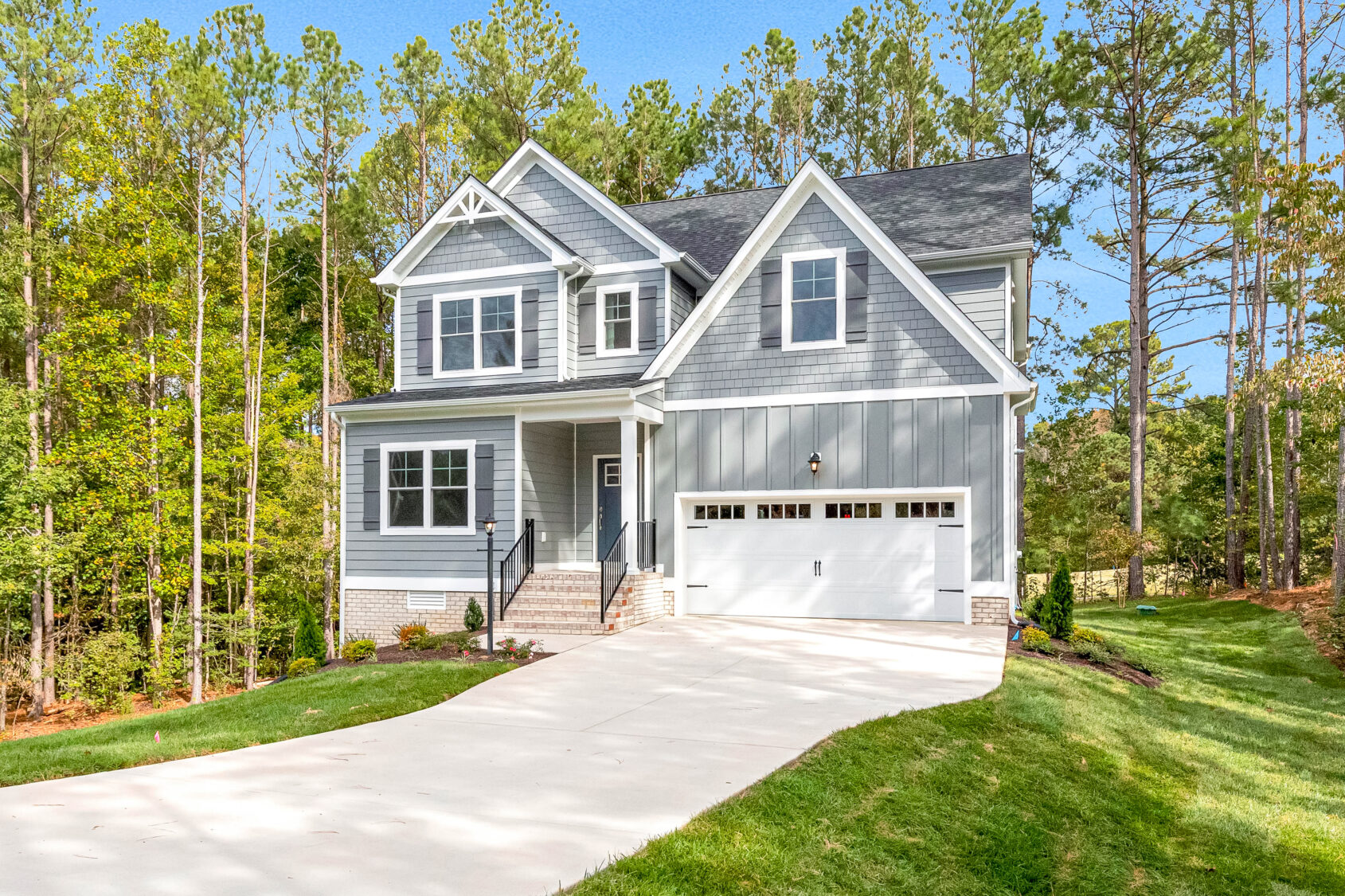 Two-story gray house with white trim, attached garage, and a large driveway surrounded by trees.