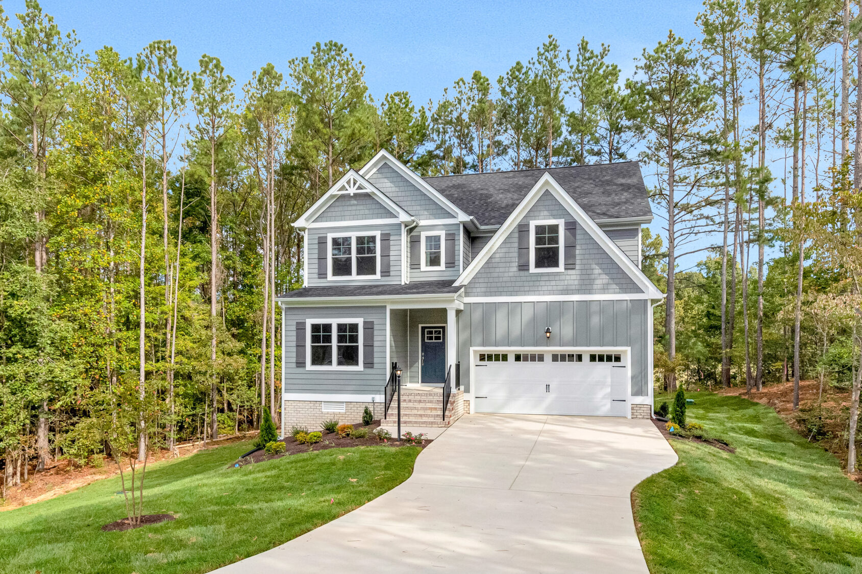 Two-story gray house with a white garage, surrounded by trees and a well-kept lawn under a clear sky.