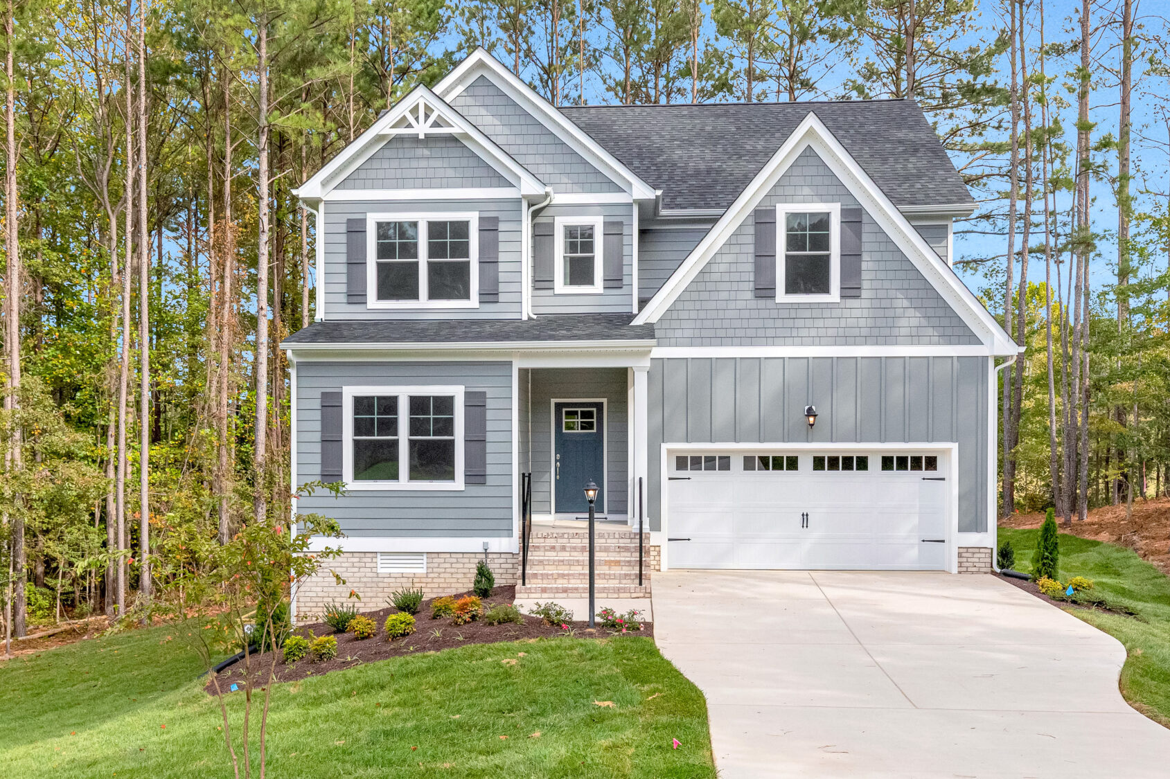 Two-story blue house with white trim, a front porch, and attached garage, surrounded by trees and greenery.