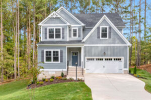 Two-story blue house with white trim, a front porch, and attached garage, surrounded by trees and greenery.