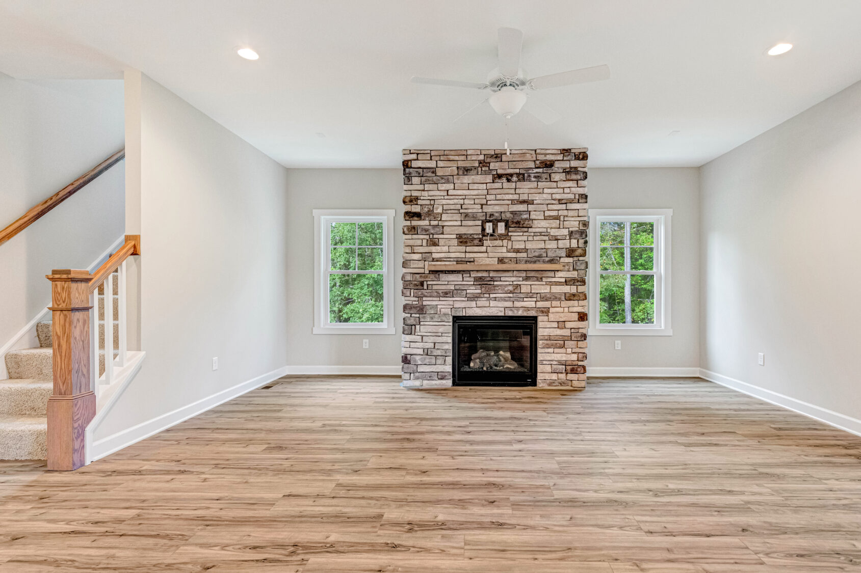 Bright living room with wood floors, stone fireplace, ceiling fan, two windows, and a carpeted staircase.