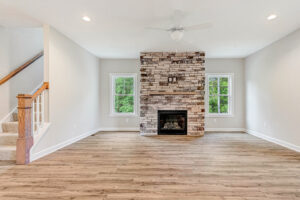 Bright living room with wood floors, stone fireplace, ceiling fan, two windows, and a carpeted staircase.