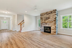 Bright living room with wood floors, a stone fireplace, and large windows, near a staircase and front door.