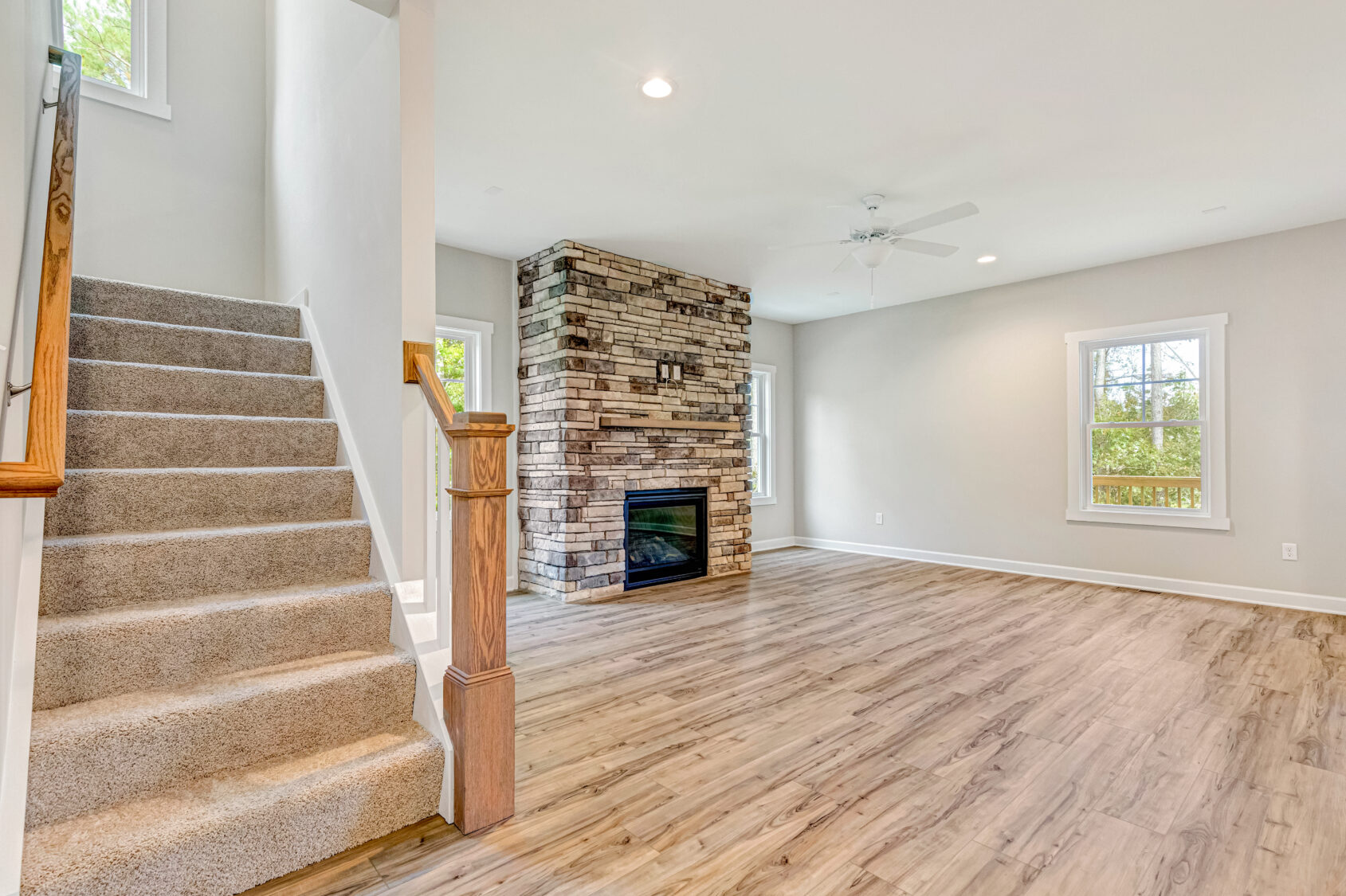 Bright living room with wood floors, carpeted stairs, and a stone fireplace against a light-colored wall.